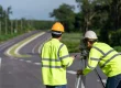 Road construction surveyors using equipment along a newly paved roadway surrounded by greenery.