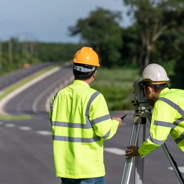 Road construction surveyors using equipment along a newly paved roadway surrounded by greenery.