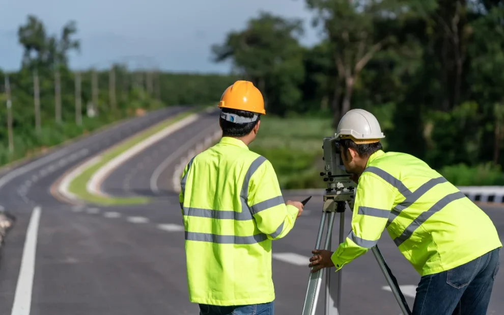 Road construction surveyors using equipment along a newly paved roadway surrounded by greenery.