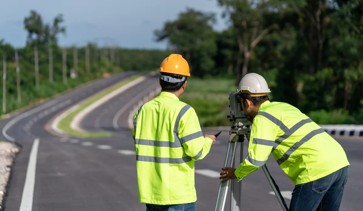 land-survey-requirements-stages-1 Road construction surveyors using equipment along a newly paved roadway surrounded by greenery.